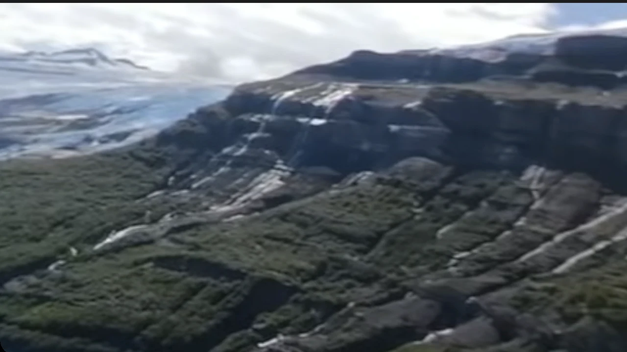 Inside a 360 degree video, looking to the side from a small plane over a valley: a glacier and snowfields above steep cliffs and green forest.